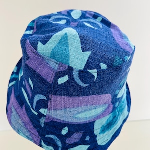 May include: A blue bucket hat featuring a repeating pattern in shades of blue and purple. The hat has a brim and a rounded crown. The hat has a tag that says "MUALA".