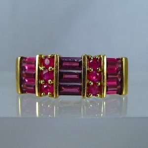 Pode incluir: Um anel de banda de ouro com uma fileira de pedras retangulares cor-de-rosa e uma fileira de pedras redondas cor-de-rosa de cada lado.
