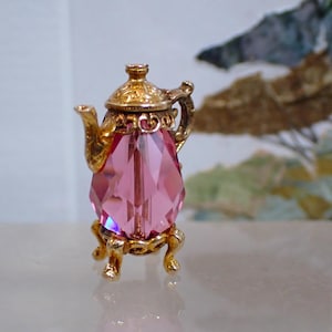 Peut inclure: Une petite théière décorative avec un corps en cristal rose facetté et des accents en métal doré orné. La théière repose sur un support doré, mettant en valeur un mélange d'élégance et de charme. Le design rappelle le décor vintage.