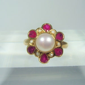 Könnte beinhalten: Ein goldener Ring mit einer zentralen Perle, umgeben von roten Edelsteinen und kleinen Perlen in einem floralen Muster. Der Ring ist vor einem neutralen Hintergrund platziert, was sein detailliertes Design und seine Handwerkskunst hervorhebt.