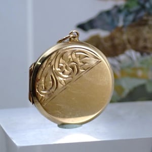 May include: A round gold-toned locket pendant featuring a polished finish on the lower half and an intricate, engraved floral pattern on the upper diagonal section. The locket includes a small circular bail at the top for attachment to a chain.
