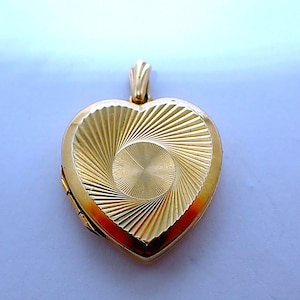 May include: A gold heart-shaped locket with a textured, radiating design on the front. The locket has a small loop at the top for a chain. The gold colour is bright and shiny, reflecting light.