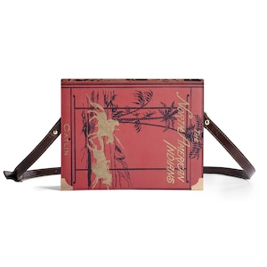 May include: A rectangular crossbody bag with a red cover featuring gold illustrations and text that reads "North American Indians". The bag has a brown strap and gold corner accents. The name "CAITLIN" is printed on the side.