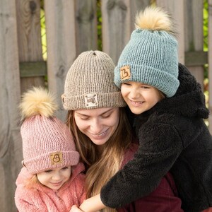 May include: Three people wearing knitted hats with pom-poms. The hats are beige, pink, and blue with brown leather patches that have letters on them. The people are smiling and hugging.