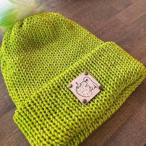 Könnte beinhalten: Hellgrüne Strickmütze mit passendem Bommel und Lederaufnäher. Der Aufnäher zeigt ein Geister- und Erdbeer-Design. Die Mütze ist aus einem dicken, strukturierten Garn gefertigt.
