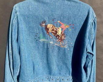 May include: Light blue denim jacket with a detailed embroidered design on the back. The embroidery depicts a rodeo scene with a cowboy on a bucking bronco. The collar and cuffs are embellished with silver studs. The jacket is hanging on a white hanger.