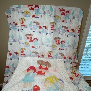 May include: A white twin-sized bed sheet and pillowcase set featuring a repeating pattern of Cabbage Patch Kids dolls in various poses. The dolls are wearing colorful clothing and are surrounded by flowers and trees. The pillowcase has a larger image of two dolls playing with a toy.