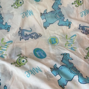 May include: A white fabric sheet featuring a repeating pattern of Monsters, Inc. characters including blue Sulley, green Mike, and a purple lizard-like creature. The print includes the text "Sulley", "Mike", "Scare Team", and the Monsters, Inc. logo.