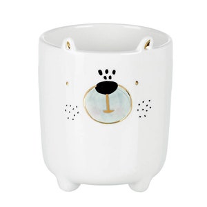 May include: White ceramic mug with a bear face design. The bear has black eyes, a black nose, and a light blue mouth with a gold outline. The mug has small feet and ears.