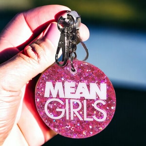 May include: Pink glitter keychain with a silver key ring and the words "MEAN GIRLS" in white lettering.