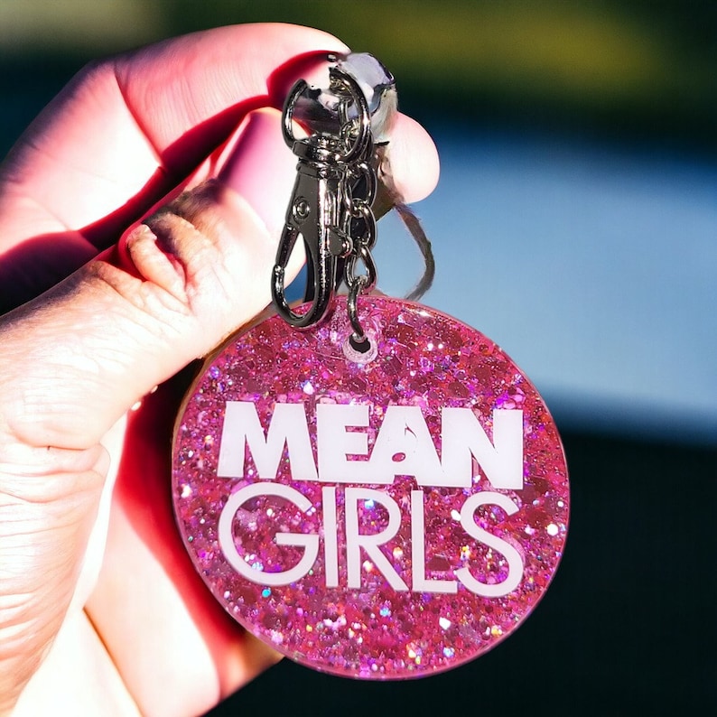 May include: Pink glitter keychain with a silver key ring and the words "MEAN GIRLS" in white lettering.