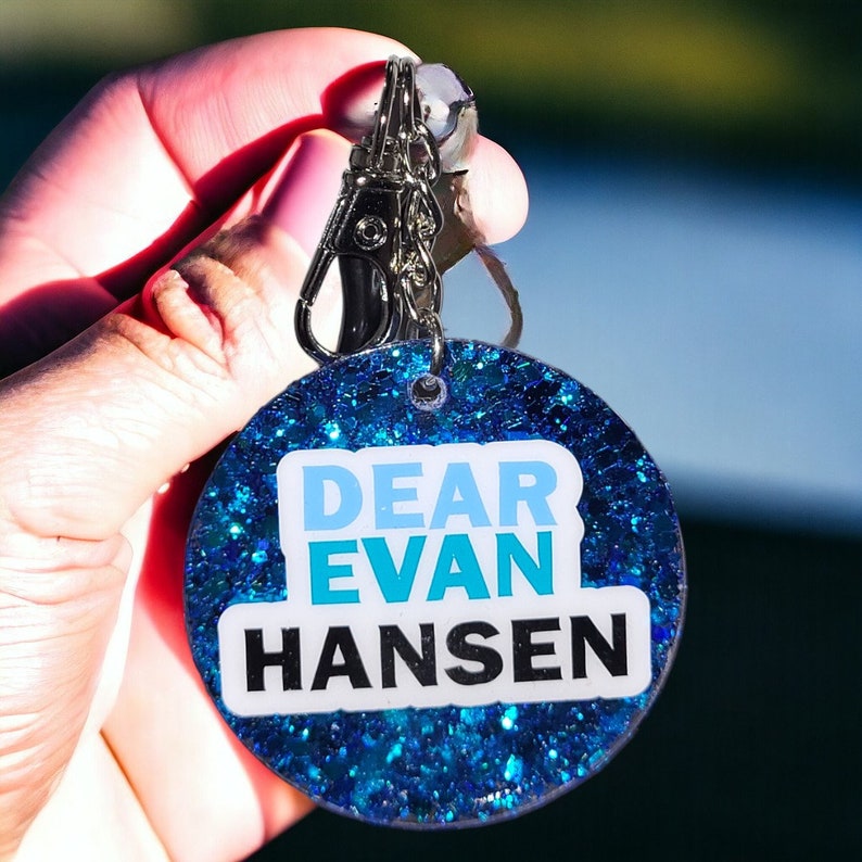 May include: A blue glitter keychain with a white sticker that says "Dear Evan Hansen".