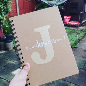 May include: A brown spiral bound notebook with a gold letter "J" and the name "Jenna" written in white cursive on the front cover.