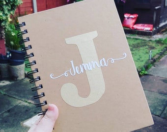 May include: A brown spiral bound notebook with a gold letter "J" and the name "Jenna" written in white cursive on the front cover.