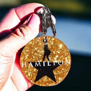 May include: A gold glitter keychain with a black silhouette of a person and the word "HAMILTON" in white letters. The keychain is attached to a silver key ring.