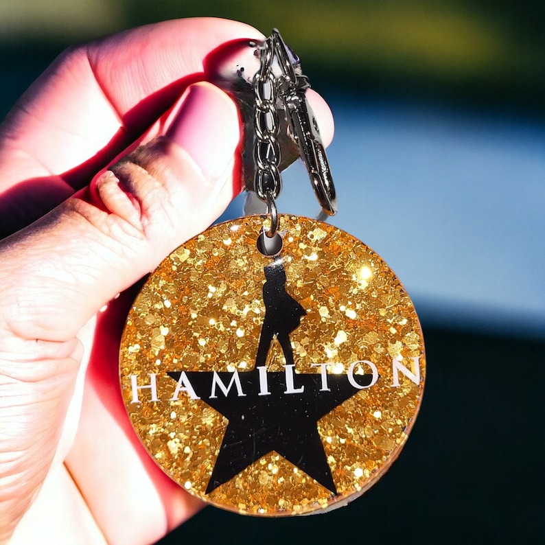 May include: A gold glitter keychain with a black silhouette of a person and the word "HAMILTON" in white letters. The keychain is attached to a silver key ring.