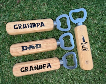May include: Wooden bottle openers with stainless steel tops. Two openers are engraved with "GRANDPA", one with "DAD" and a mustache, and one with "DADDY NEED BEER" and a bottle graphic. The openers are on a green grassy surface.