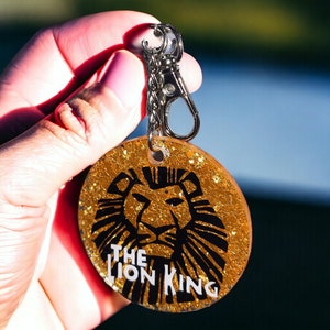 May include: A gold glitter keychain with a black silhouette of a lion's head and the text "The Lion King".