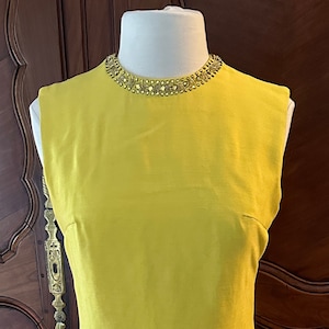 May include: A sleeveless yellow maxi dress with a beaded neckline. The dress is on a mannequin and is displayed against a wooden cabinet.