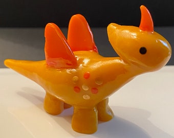 May include: A small, yellow ceramic dinosaur with orange spikes and gold accents. The dinosaur has a small horn on its head and a simple black eye.