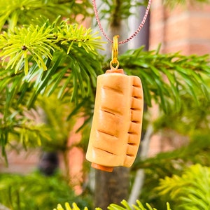 May include: A light brown pendant shaped like a hot dog in a bun, suspended from a gold-coloured hoop and a pink string. The pendant features carved details to resemble the bun and hot dog. The background is a blurred view of green foliage.