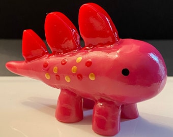 May include: A pink ceramic dinosaur figurine with gold and red spots. The dinosaur has a long tail, four legs, and three spines on its back.