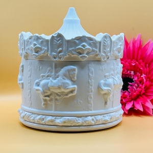 May include: White ceramic carousel music box with a detailed design of horses and a roof. The box is closed and sitting on a light brown surface.