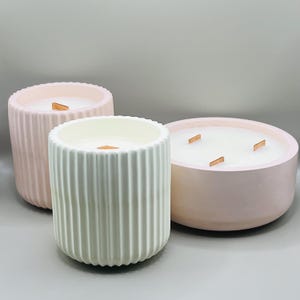 May include: Three candles in different sizes, all with a white wax and wooden wicks. The candles are in pink and white concrete containers with a ribbed texture.