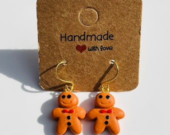 May include: A pair of handmade gingerbread man earrings with gold-colored hooks. The gingerbread men are brown with black eyes, a red bow tie, and a smiling face. The earrings are displayed on a brown card with the text "Handmade with love".