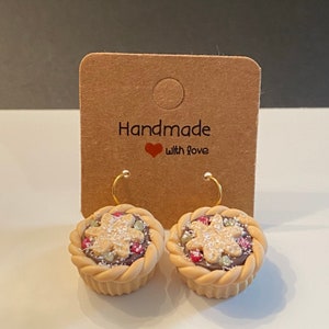 May include: A pair of handmade earrings featuring miniature pie designs with a brown crust, red and white filling, and a sprinkle of white powder. The earrings are on a brown card with the text "Handmade with love".