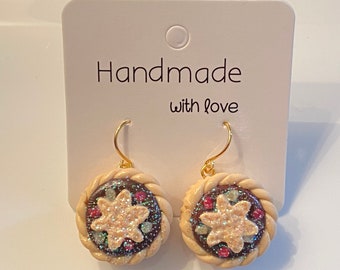 May include: A pair of handmade earrings with a circular design. The earrings feature a tan-colored outer ring with a braided texture, surrounding a dark brown center with a glittery star and colorful accents. The card behind the earrings says "Handmade with love."