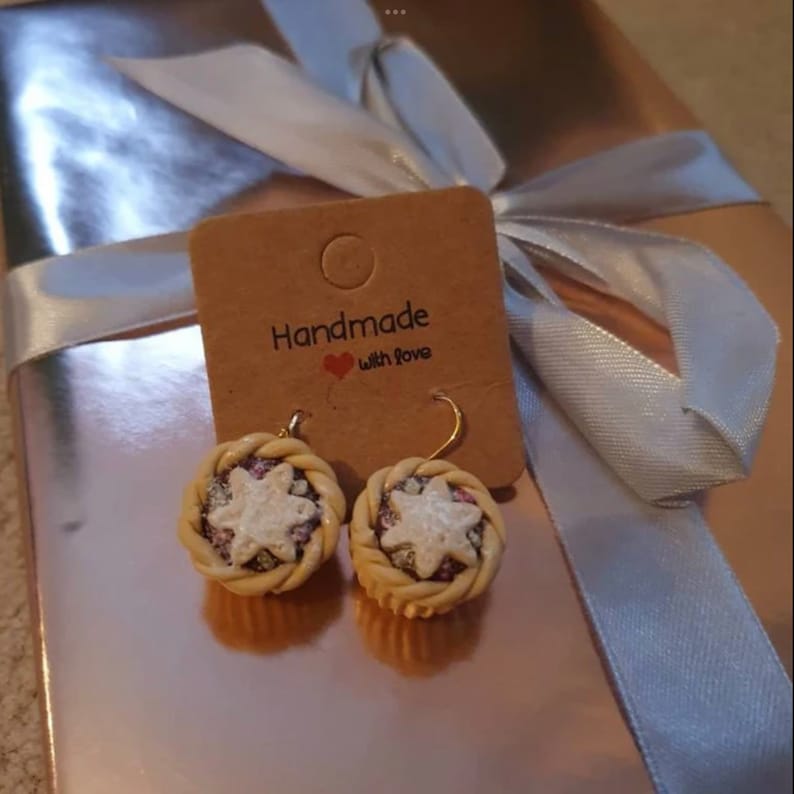 May include: A pair of handmade earrings featuring a miniature pie design with a white star and pink and white sprinkles. The earrings are on a brown card with the text "Handmade with Love".