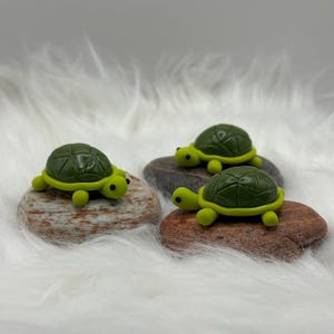 Puede incluir: Tres figuras de tortugas en miniatura. Cada tortuga tiene un caparazón verde oscuro y un cuerpo y extremidades verde claro. Las tortugas descansan sobre pequeñas piedras lisas y multicolores. El fondo es un material suave, blanco y esponjoso.