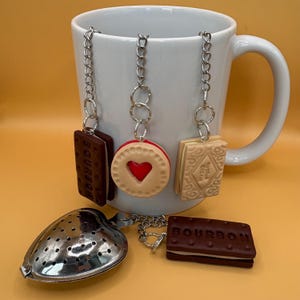 May include: A white ceramic mug featuring three hanging biscuit-shaped charms on silver chains: a dark brown Bourbon biscuit, a round beige biscuit with a red heart centre, and a light beige Custard Cream biscuit. A heart-shaped tea infuser is included.