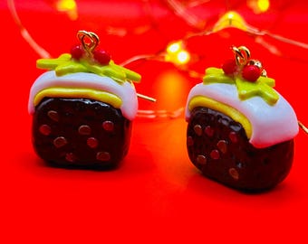 May include: Two Christmas pudding earrings. Each earring features a dark brown, cube-shaped base with red dots, topped with white icing, yellow cream, green holly, and red berries. The earrings have gold-colored hooks and are set against a red background.