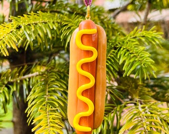 May include: A hot dog ornament with a brown bun, a reddish-brown sausage, and a yellow mustard squiggle. The ornament hangs from a purple ribbon and gold-colored metal loop. The background is a green, leafy tree.