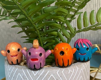 May include: Four small ceramic ghost figurines with different colors and facial expressions. The ghosts are orange, purple, blue, and gold. They are sitting on a gray and white patterned surface.