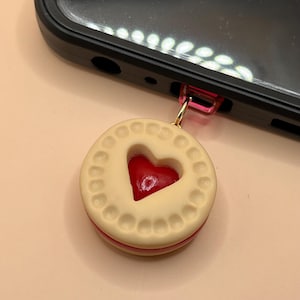May include: A small, white cookie with a red heart-shaped filling. The cookie is attached to a gold chain and a pink phone charm.