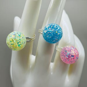 May include: Three silver rings with iridescent glitter balls. The balls are light blue, light green, and pink.