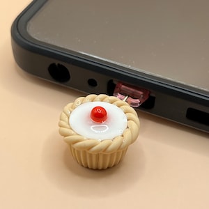 May include: A small, beige cupcake with a white frosting and a red cherry on top. The cupcake is attached to a pink and clear plastic connector.