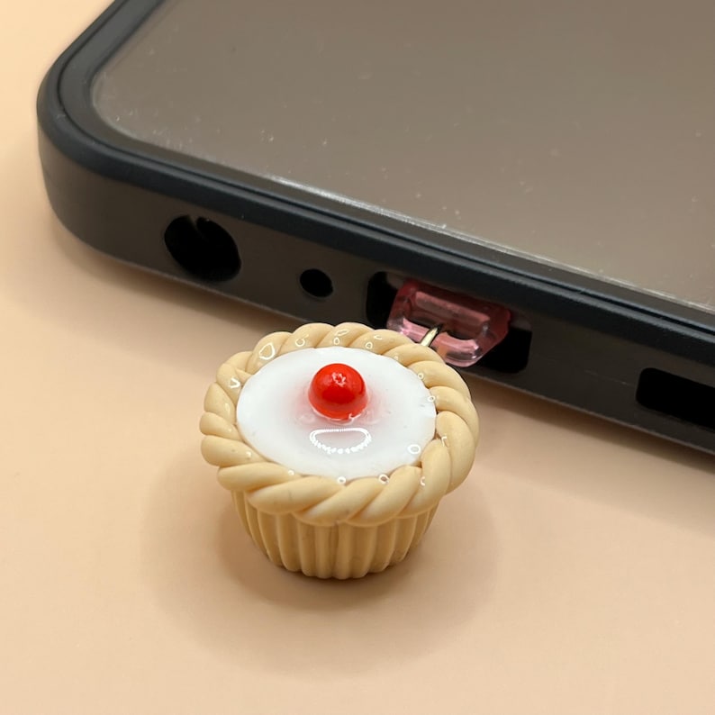 May include: A small, beige cupcake with a white frosting and a red cherry on top. The cupcake is attached to a pink and clear plastic connector.