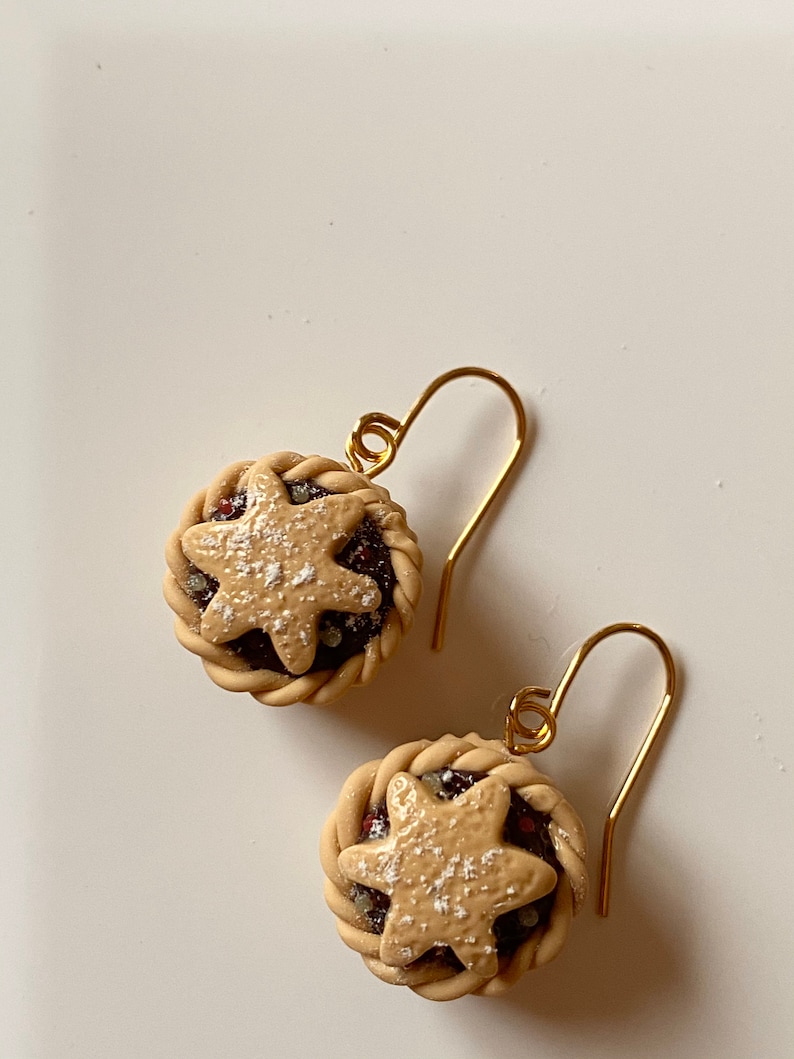 May include: A pair of gold dangle earrings featuring miniature pie crusts with a star-shaped design and a sprinkle of powdered sugar.