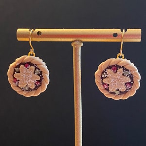 May include: A pair of gold dangle earrings featuring a miniature pie design. The pie is made of polymer clay and has a brown crust, a red and white filling, and a white sugar sprinkle topping.