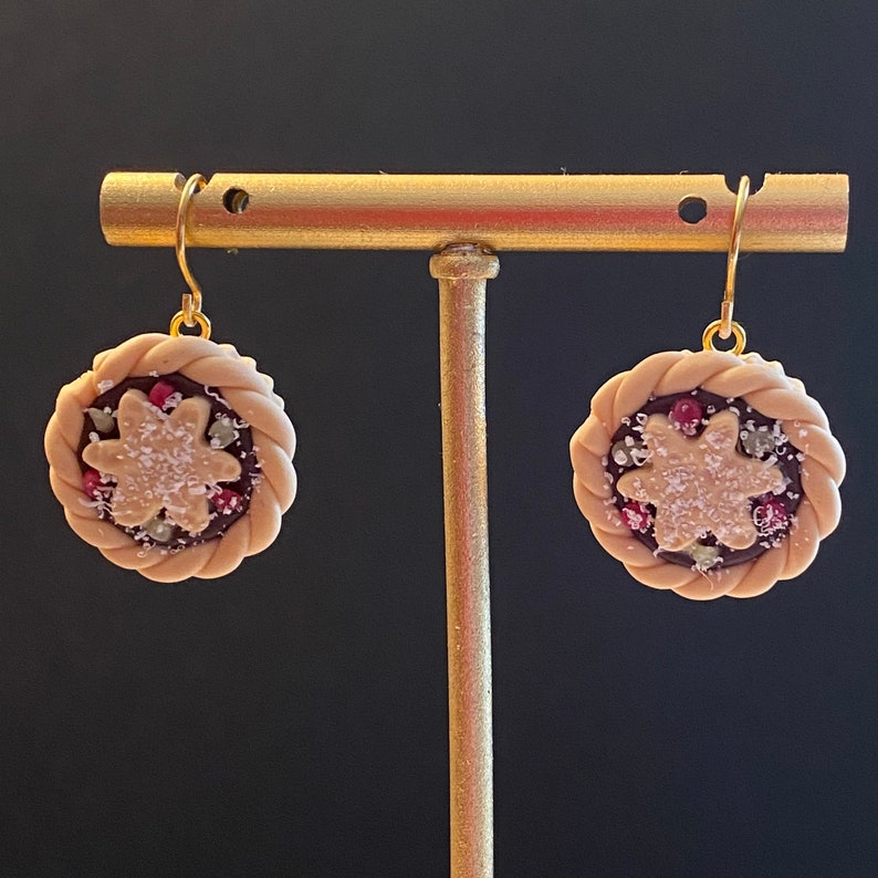 May include: A pair of gold dangle earrings featuring a miniature pie design. The pie is made of polymer clay and has a brown crust, a red and white filling, and a white sugar sprinkle topping.