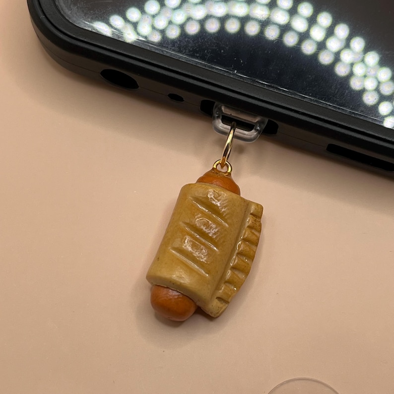 May include: A small, brown, and orange,  miniature hot dog charm with a gold chain. The charm is attached to a black cell phone case.
