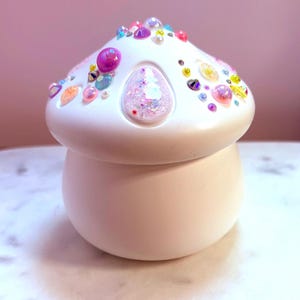 May include: A white mushroom-shaped trinket box with a rounded base and a decorative top. The top is adorned with colorful rhinestones, gems, and pearls. The box has a smooth, matte finish and a whimsical design, perfect for storing small items.