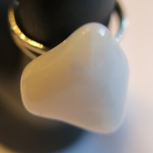 May include: A close-up of a ring featuring a light-colored, smooth, rounded stone. The ring's band is a thin, shiny gold color. The stone is set against a dark, blurred background, highlighting the ring's design.