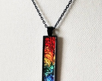 May include: A rectangular pendant necklace featuring a vibrant, multi-coloured foil insert with a crinkled texture. The pendant is encased in a thin black metal frame and hangs from a matching black metal chain, displaying a spectrum of colours.