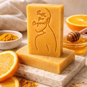 Peut inclure: Gros plan sur trois savons rectangulaires de couleur orange. Le savon du dessus porte un motif en relief avec le texte "Soothe the Organics". Des tranches d'oranges, de la poudre de curcuma et du miel sont également visibles.