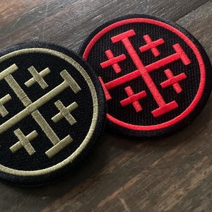 Op de afbeelding: Twee ronde geborduurde patches. Eén is zwart met een gouden ontwerp, de andere is zwart met een rood ontwerp. Beide hebben een kruisachtig symbool binnen een cirkelvormige rand. De patches liggen op een houten oppervlak.